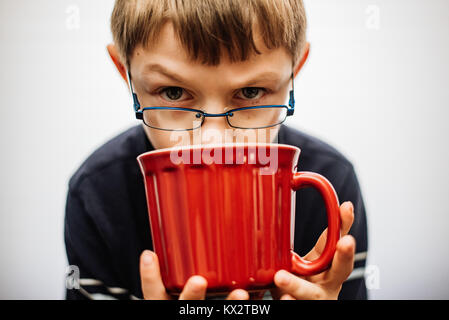 Ein 11 Jahre alter Junge Brille über den Rand eine rote Tasse Tee suchen. Stockfoto