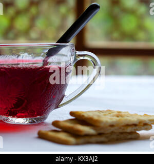 Selektiver Fokus auf transparent Tasse Hibiskus Tee auf weißen Tisch Fenster gegen Fenster Hintergrund und serviert mit Cracker - Seitenansicht Stockfoto