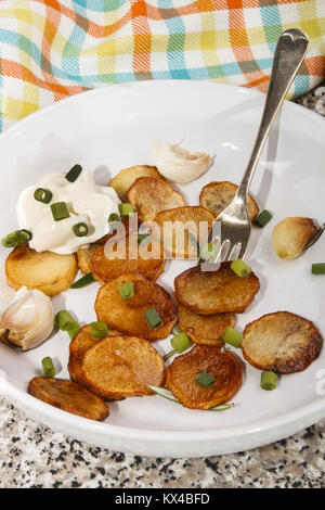 Gebratene Kartoffelscheiben mit Frühlingszwiebeln, saure Sahne und Rosmarin auf eine tiefe weiße Platte Stockfoto