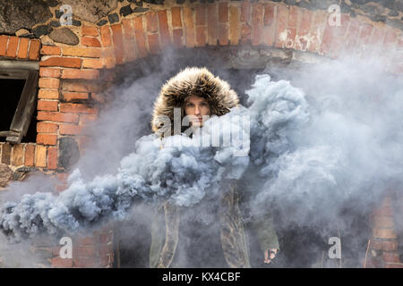 Jungen kaukasischen Mann in einem grünen Winter parka posiert mit einem grauen Rauch Bombe auf einem düsteren Wintertag. Tartu, Estland. Stockfoto