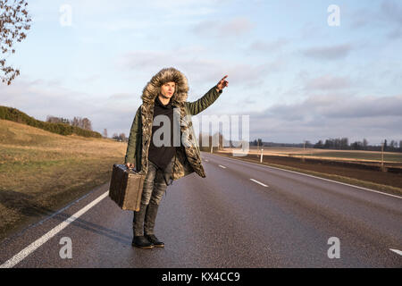 Jungen kaukasischen Mann in einem warmen Winter parka hageln ein Taxi auf eine Landstraße mit einem Vintage Koffer. Tartu, Estland. Stockfoto