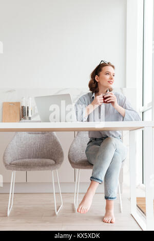 Portrait von Fröhliche Frau in gestreiften Hemd und Jeans holding Tasse Kaffee beim Sitzen auf weichen Stuhl in der Küche Stockfoto