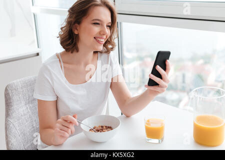 Portrait von Süß lächelnde Mädchen in weißen T-Shirt plaudern auf Smartphone beim Sitzen und Essen Cornflakes am Küchentisch Stockfoto