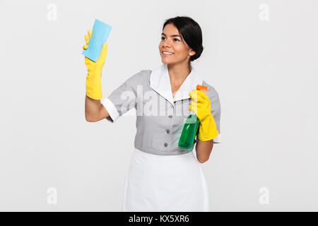 Porträt einer fröhlichen hübsche Haushälterin in Uniform Reinigung mit Schwamm und Feldspritze gekleidet beim Stehen auf weißem Hintergrund Stockfoto