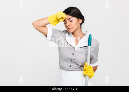 Nahaufnahme von Müde brunette Mädchen in Uniform holding Mop und wischt sich den Schweiß von der Stirn, auf weißem Hintergrund Stockfoto