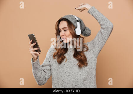 Junge Frau mit Curly Frisur holding Smartphone niedlich, während Sie Musik hören, auf beige Hintergrund isoliert Stockfoto