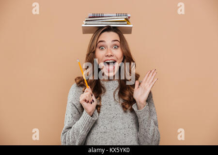 Portrait von überglücklich Junge brunette Mädchen in grau Jersey holding Bücher auf dem Kopf, an der Kamera suchen, über beigen Hintergrund isoliert Stockfoto