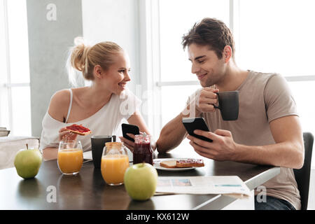 Gesunde junge lächelnde Paar mit ihren Smartphones und Reden beim Essen in der Küche das Frühstück Stockfoto