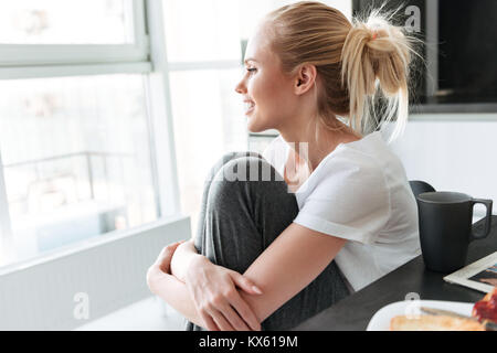 Seitenansicht von der hübschen blonden Dame zur Seite schauen und lächeln, während in der Küche sitzt Stockfoto