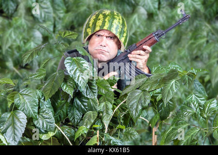 Lustige Soldat mit einem Maschinengewehr AK47 aus der grünen Blättern. Crazy man in einen Helm der Wassermelone patrouillieren mit Kalaschnikow in der Hand. Stockfoto