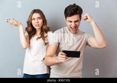 Porträt eines jungen Paares mit Handy, man spielt Spiele auf Handy während frustrierte Mädchen stand in der Nähe und Suchen an Kamera Stockfoto