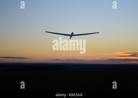 Ein Segelflugzeug wird in den Sonnenuntergang gestartet, mit dunklem Boden unten und dem roten Himmel am Horizont, mit ein paar Wolken. Der Schirm steigt nach oben in die Luft. Stockfoto