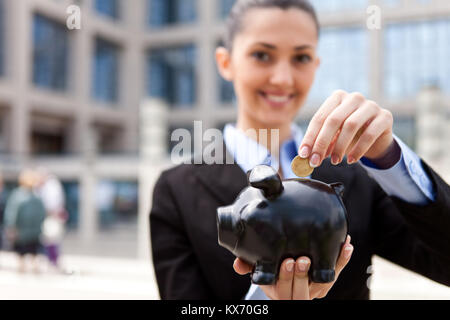 Schöne junge Frau, eine Münze in ein großes rosa Sparschwein vor ihrem Büro Gebäude (Fokus auf das Sparschwein) Stockfoto