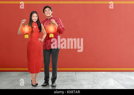 Junge chinesische Paar mit traditionellen Kleid mit roten Laternen auf rote Wand Hintergrund. Happy Chinese New Year Stockfoto