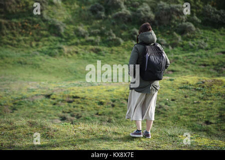 Eine Junge kaukasier Frau stehend auf einem grasbewachsenen Hügel, indem sie sich mit einem Rucksack und einem langen Rock auf dem Schauen in die Ferne bereit, die Lan zu erkunden Stockfoto