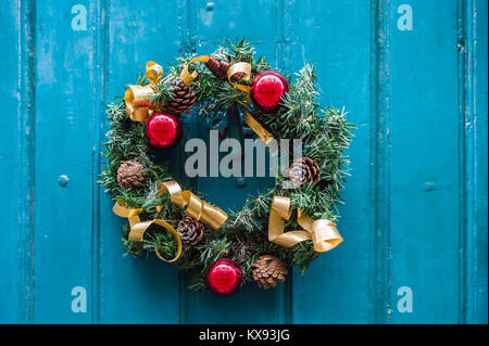 Vorderansicht eines traditionellen Weihnachtskranzes aus Kiefernästen, Tannenzapfen, roten Weihnachtskugeln und goldenen Bändern an einer alten Holztür Stockfoto