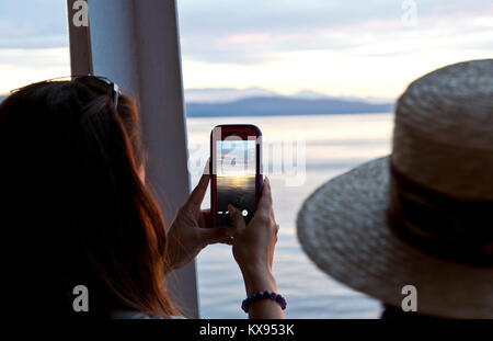 Frau ein Foto mit Ihrem Handy auf den Sonnenuntergang auf dem Wasser. Auf der BC-Fähre auf der Straße von Georgia. Stockfoto