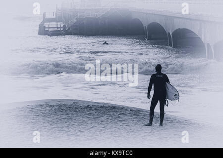 Surfer auf dem Strand in der Nähe von Bournemouth Pier mit Blick auf das Meer als Surfer die Brandung an einem windigen Tag am Strand von Bournemouth, Dorset im Januar Stockfoto