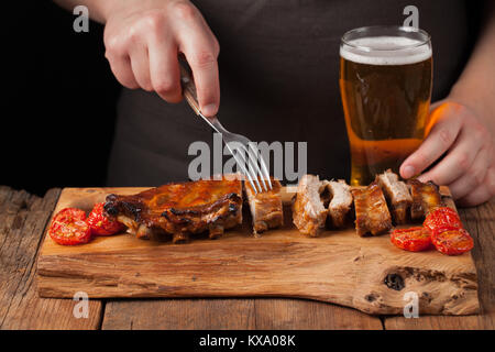 Männer Koch Spieße mit einer Gabel bereit Rippchen essen, liegend auf einem alten Holztisch. Ein Mann versucht, leichte Snacks und Getränke Bier auf schwarzem Hintergrund. Stockfoto