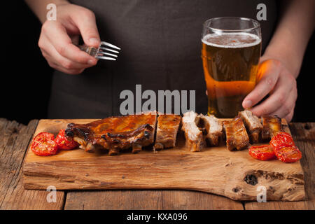 Männer Koch Spieße mit einer Gabel bereit Rippchen essen, liegend auf einem alten Holztisch. Ein Mann versucht, leichte Snacks und Getränke Bier auf schwarzem Hintergrund. Stockfoto