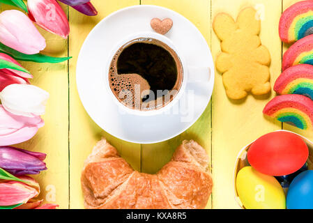 Ostern Morgen mit einer Tasse Kaffee, Regenbogen cookies, bemalte Eier, Häschen geformte Cookie, Croissant und vielfarbigen Tulpen, gelb Holztisch. Stockfoto