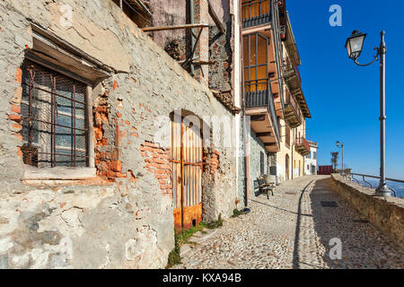 Schmale Straße mit Kopfsteinpflaster entlang der alten Häuser und Laternen unter blauem Himmel in der kleinen Stadt von La Morra in Piemont, Norditalien. Stockfoto