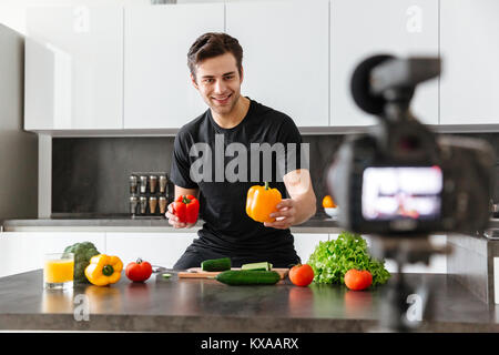 Fröhlicher junger Mann seinem Video Blog Episode über gesundes Essen kochen nehmen, während in der Küche stehend Stockfoto