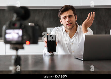Hübscher junger Mann nimmt sein Video Blog Episode über die neuen technischen Geräte während am Küchentisch sitzen und mit leerer Bildschirm Handy Stockfoto