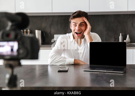 Glückliche junge Mann nimmt sein Video Blog Episode über die neuen technischen Geräte während am Küchentisch sitzen mit leerer Bildschirm Laptop Stockfoto