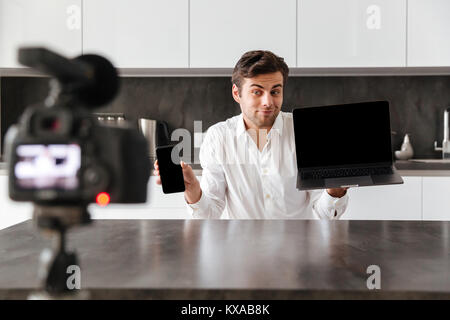 Lächelnden jungen Mann nimmt sein Video Blog Episode über neue technische Geräte und zeigt leeren Bildschirm Handy und Laptop zuhause Stockfoto