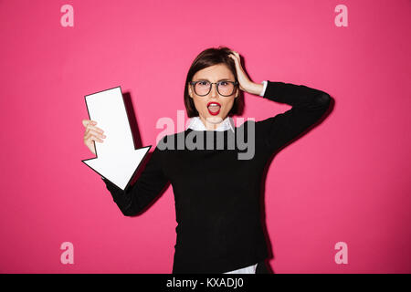 Porträt von einem aufgeregten Jungen Geschäftsfrau holding Papier Pfeil nach unten über rosa Hintergrund isoliert Stockfoto