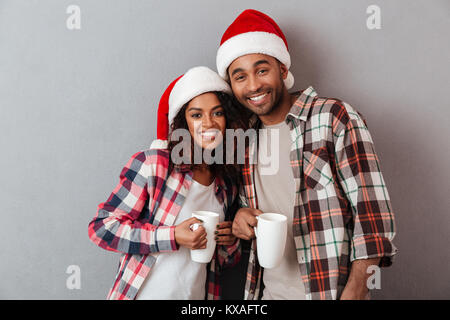 Portrait eines glücklichen jungen afrikanischen Paar in Weihnachten hüte Umarmen und Halten Sie Tassen mit Tee über grauen Hintergrund gekleidet Stockfoto