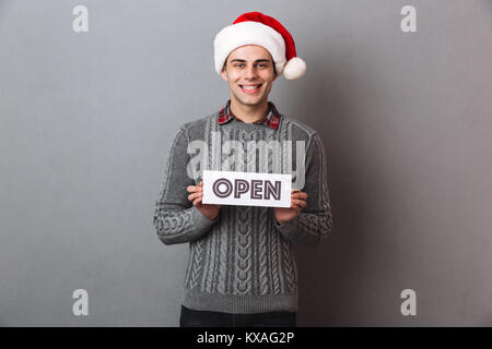 Image der Jungen fröhlichen Mann, weihnachten santa hut stehend über graue Wand, Papier mit Open Text isoliert. Suchen Kamera. Stockfoto