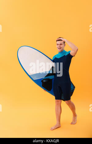 Volle Länge Bild der junge lächelnde Surfer gehen mit Surfbrett und entfernten Blick auf gelbem Hintergrund Stockfoto