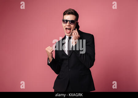 Bild der glückliche junge Unternehmer stehen isoliert mit Sonnenbrille und Gespräch per Handy Sieger Geste machen. Stockfoto