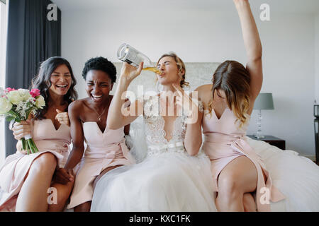 Braut im weißen Kleid trinken eine Flasche Wein beim Sitzen auf dem Bett mit Brautjungfern. Braut und Brautjungfern genießen vor der Hochzeit im Hotel Zimmer. Stockfoto