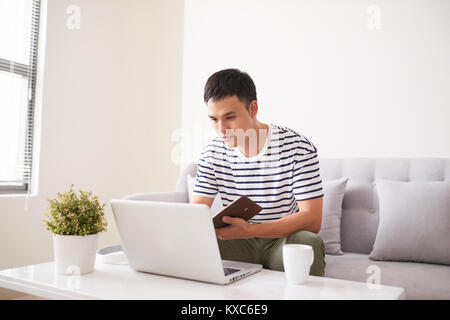 Stattliche ernster Mann, am Laptop Bildschirm schaut und etwas in sein Notizbuch, sitzen auf der Couch Stockfoto