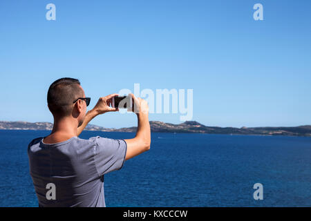 Nahaufnahme eines jungen kaukasischen Mann von hinten nehmen Sie ein Foto auf das Meer an der Costa Smeralda, Sardinien, Italien gesehen, mit seinem Smartphone, mit einem Bla Stockfoto