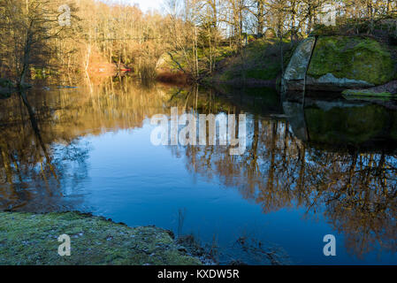 Die sanften Strömung eines Flusses. Sunshine hits den Wald im Hintergrund. Moos bedeckt, gerissene Boulder am Flussufer. Kleine Verwirbelungen im Wasser. Die Stockfoto