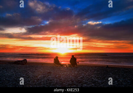 Sonnenuntergang bei Gillespies Strand in Fox Glacier, West Coast, Neuseeland. Zwei Reisende - Mann und Frau Silhouetten entspannen auf Camping Stuhl und beobachten Stockfoto