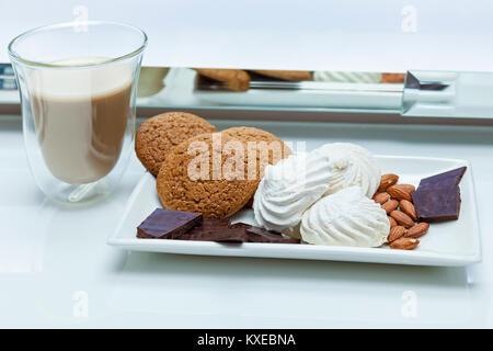 Ein Glas Kaffee mit Gebäck Brocken der Schokolade, Marshmallows und Muttern auf einem weißen Tisch. Ein Glas Kaffee mit Süßigkeiten Stockfoto