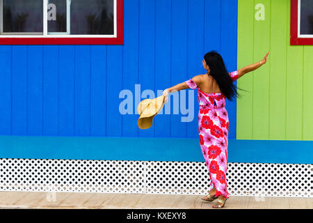 Ziemlich polynesische Inselbewohner in einem roten Kleid mit Blumenmuster und Sonnenhut tanzen vor einem bunten Grün und Blau Holzwand in Hawaii Stockfoto