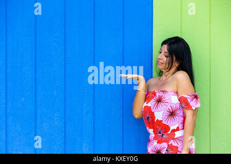 Junge Frau an Ihrer leeren Hand auf der Suche nach Product Placement erweitert, als sie in einem roten Blumen Sommer Kleid vor einem blauen und grünen Holz wa steht Stockfoto