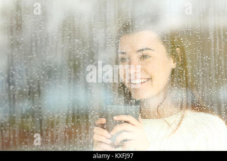 Positive glücklich jugendlich durch ein Fenster mit einer Kaffeetasse in der einen regnerischen Tag suchen Stockfoto