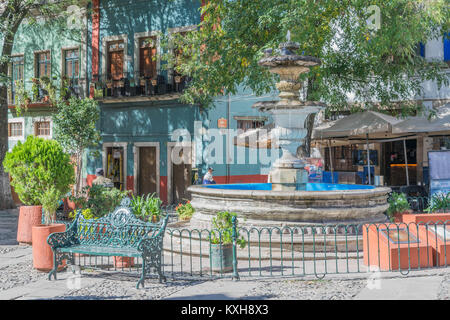 Zentrum Brunnen, eine grüne eisen Bank, etwas Grün, Schirme für Speisen und bunte 2-stöckigen Gebäuden, die in der San Fernando Plaza, in Guanajuato Stockfoto