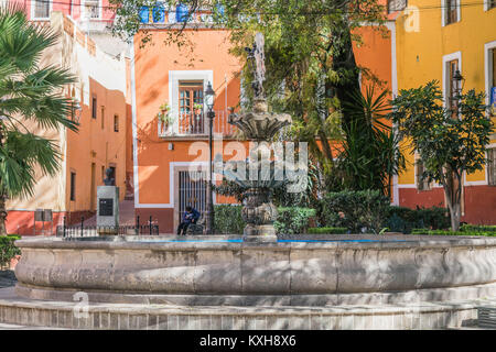 Stein Wasser Brunnen mit einem blauen Interieur, bunten Gebäude, etwas Grün und eine Person, die auf einer Bank sitzen, im Jardin de Reforma, in Guanajuato Stockfoto