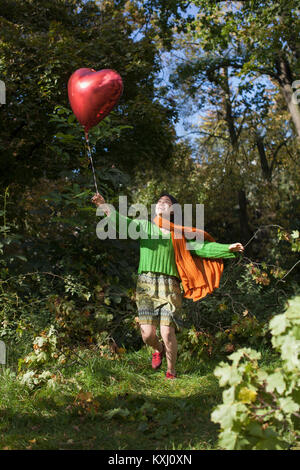 Glückliche junge Frau hält einen roten Herzballon, während sie im Park gegen Bäume läuft Stockfoto