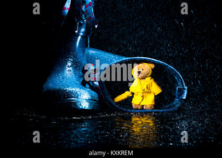 Wenig Teddybär in der gelben Regenmantel holding Umbrella finden Zuflucht in einem blauen wellie. Stockfoto