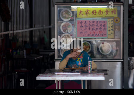 Ältere asiatische Frau saß an chinesischen Hawker restaurant Tisch Lesen mit Softdrink mit hawker im Hintergrund stand. Stockfoto