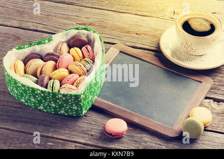 Bunte Makronen in Valentines Tag herzförmigen Geschenkbox und Kaffee Tasse auf hölzernen Tisch. Mit Tafel für Ihre Grußansagen Stockfoto